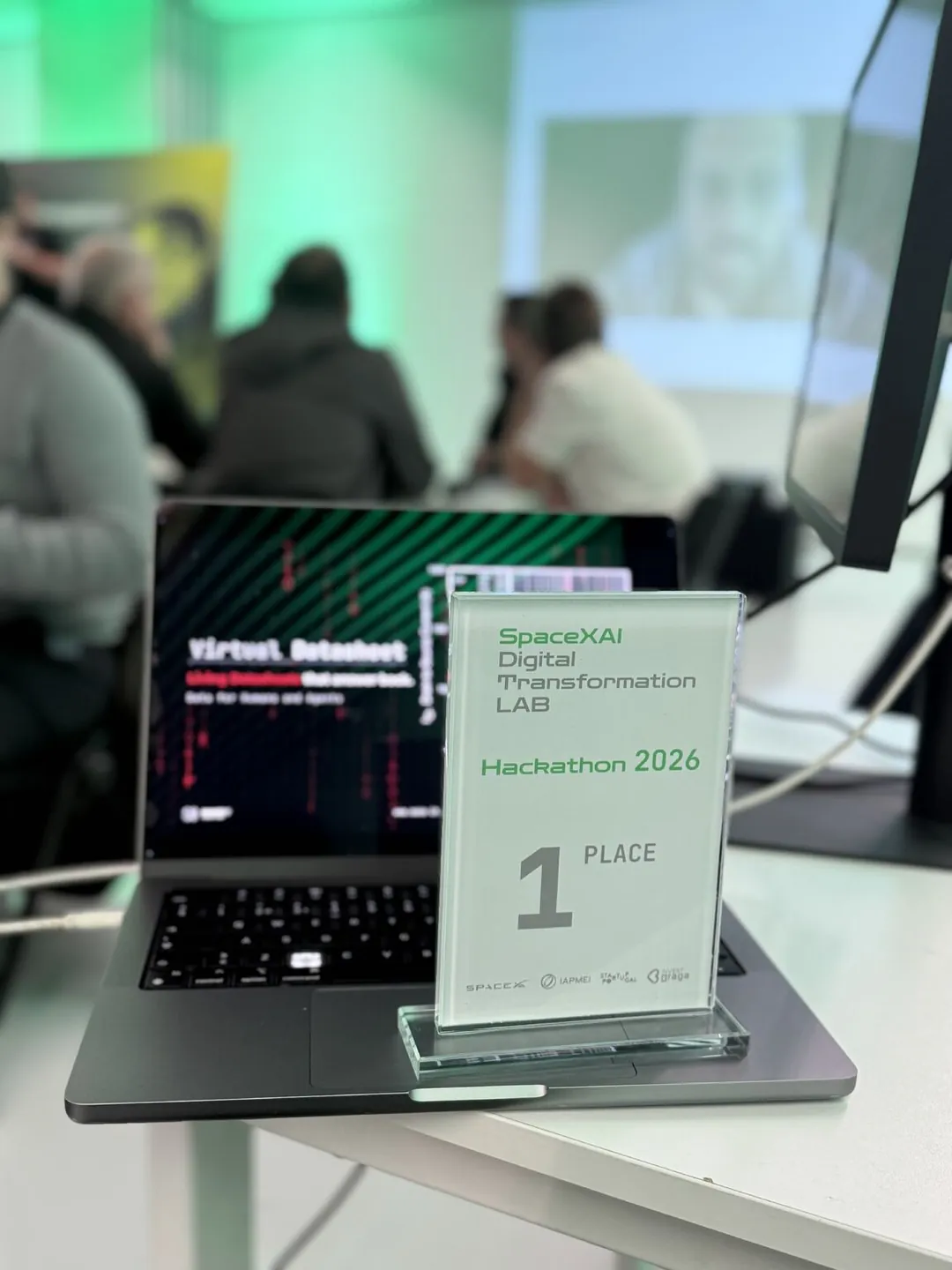 Troféu acrílico do 1.º lugar do hackathon SpaceXAI Digital Transformation Lab em frente a um portátil com o Virtual Datasheet
